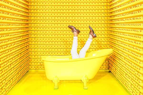 Photo of a room paneled in yellow, rubber duckies. In the middle is a yellow bathtub with legs waving in t