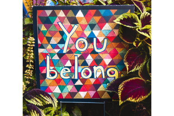 A multi-colored illustration, like a patchwork quilt, with the words "You belong" on it surrounded by plants.