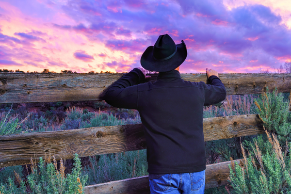 Photo of a man wearing a black cowboy hat, jacket & blue jeans. He leans against a fence & looks at a purple sky