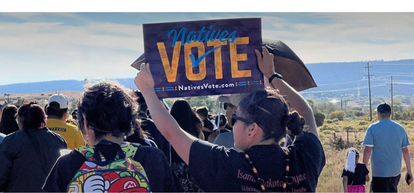 An Indigenous person holds a "Natives Vote" sign in a crowd of people with a ridge of hills in the background