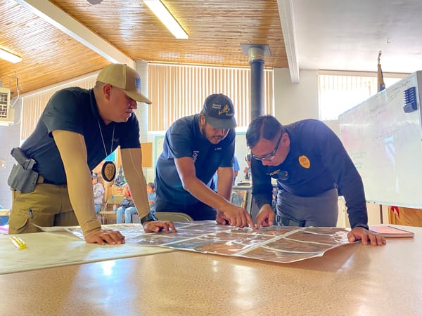 3 men wearing badges bend over a table scanning documents and maps