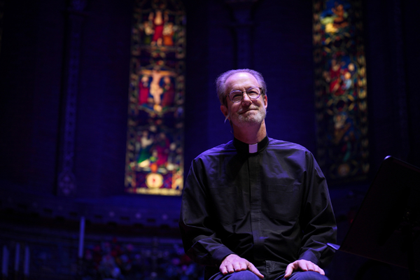 A balding, White priest wearing glassed sits in front of stained glass windows smiling