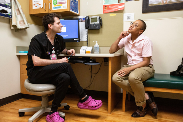 A white man in black scrubs & pink Crocs sits across from a Black Trans man in a pink shirt & khaki pants 