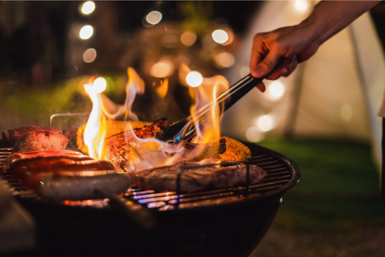 A hand uses tongs to turn meat on a flaming grill