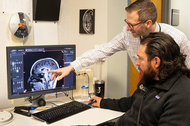 2 men look at a computer monitor displaying a brain scan as the man with glasses points at it