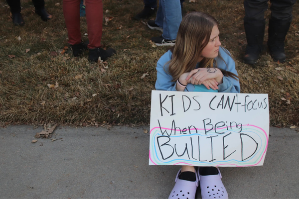 A white teen with long, brown hair sits on the grass holding a sign: "Kids can't focus when being bullied."