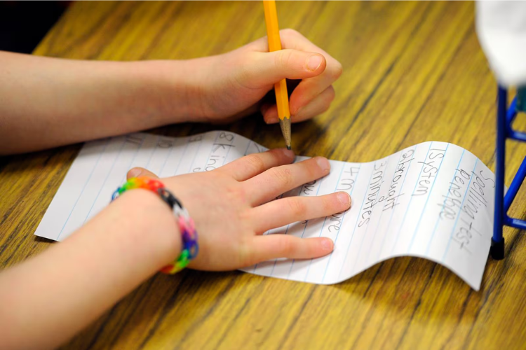 Photo of a young student's hands writing notes; on one wrist is a multi-colored band